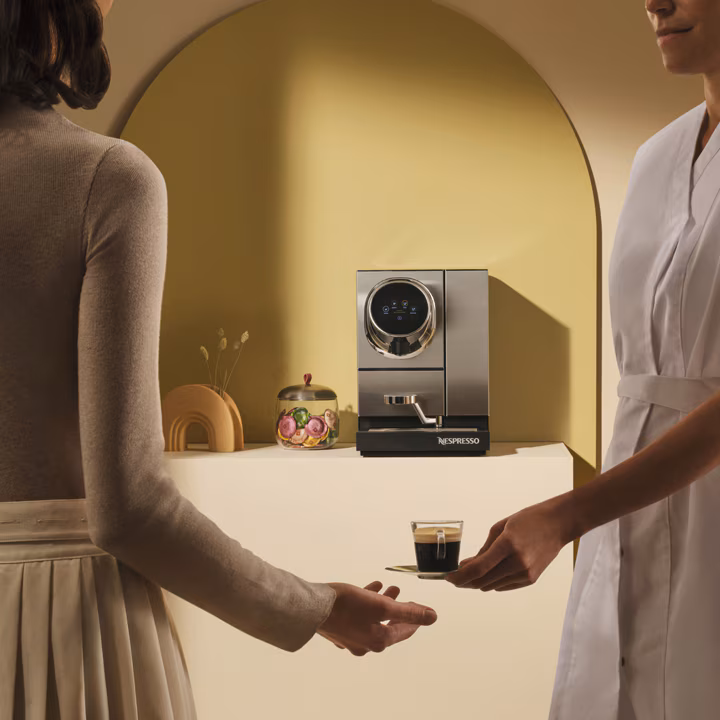 Egy férfi egy csésze espressot nyújt kollegájának, háttérben a Momento 120 kávégép látható.